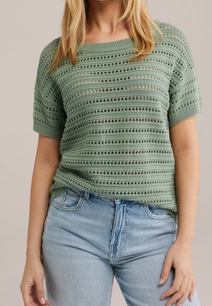 Femme portant un haut en maille vert clair à manches courtes avec des motifs ajourés horizontaux et un jean taille haute bleu clair, debout devant un fond neutre.