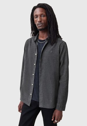 Chemise boutonnée gris foncé avec une texture subtile et un petit logo sur la poitrine, portée sur un T-shirt noir, associée à un pantalon noir.