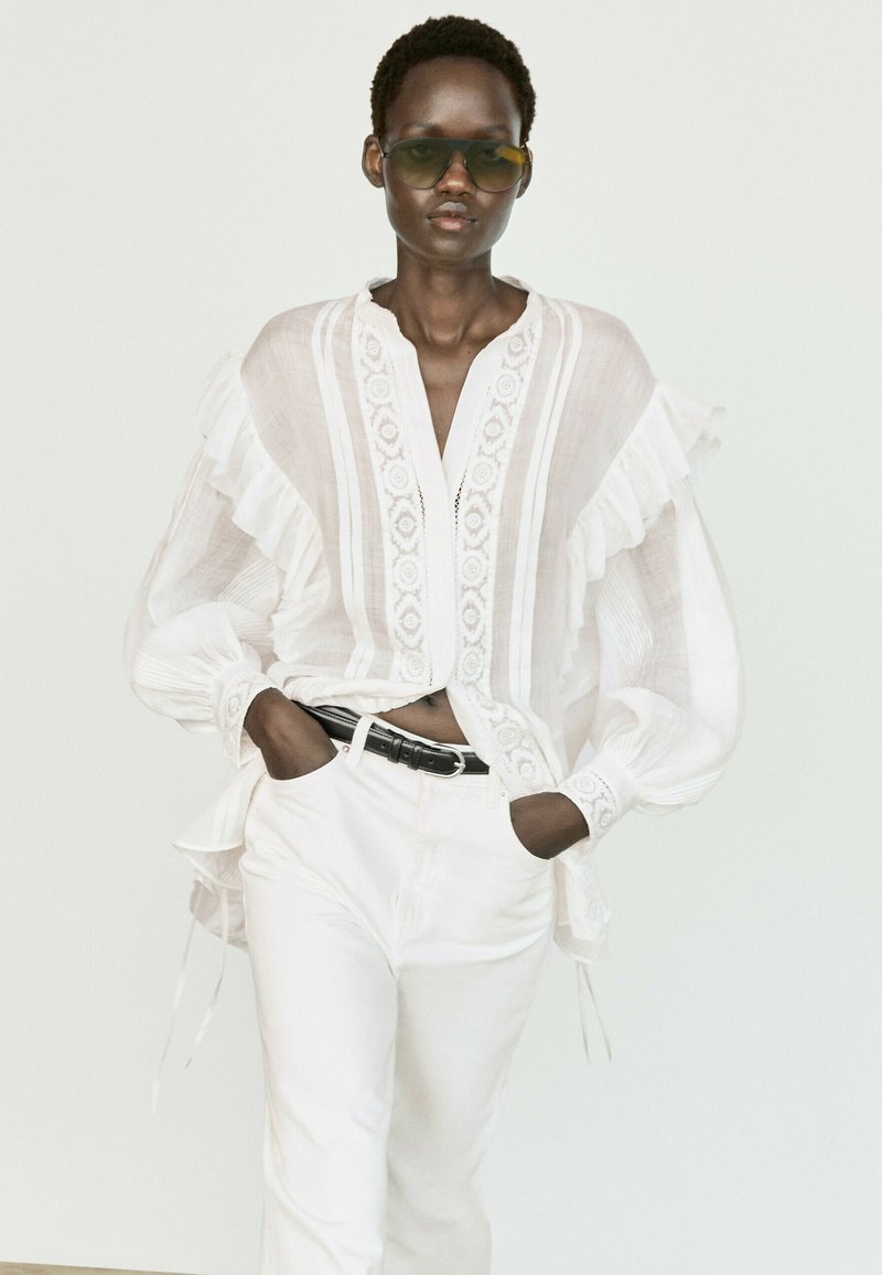 Vrouw draagt doorschijnende witte blouse met ruches, witte broek, zwarte riem en donkere zonnebril, staat met handen in zakken tegen een witte achtergrond.