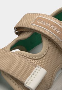 Μπεζ σανδάλι Calvin Klein με υφασμένες λουριά, κλείσιμο με βέλκρο και πράσινη εσωτερική σόλα ορατή μέσα από τμήματα με κοψίματα.