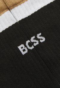 Chaussettes noires avec une texture côtelée, ornées de rayures horizontales blanches et marron. Le mot "BOSS" est tricoté en blanc sur la zone noire.