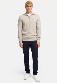 Vaaleanharmaa puolivetoketjullinen neulepaita, jossa on ribbineulosta, päällä valkoinen paita, yhdistetty tummansinisiin housuihin ja valkoisiin lenkkareihin.