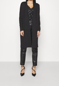 Svart cardigan med öppen front, långa ärmar och grovstickad struktur. Kombinerad med en blus med prickar och figurnära byxor i konstläder. Svarta klackar.