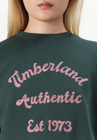 Zelená mikina s ružovým vyšitým textom: "Timberland Authentic Est 1973." Hladká tkanina s dizajnom okrúhleho výstrihu.