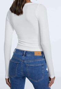 Frau in einem enganliegenden weißen Langarmoberteil und blauen Jeans, von hinten vor einem einfarbigen hellen Hintergrund gezeigt.