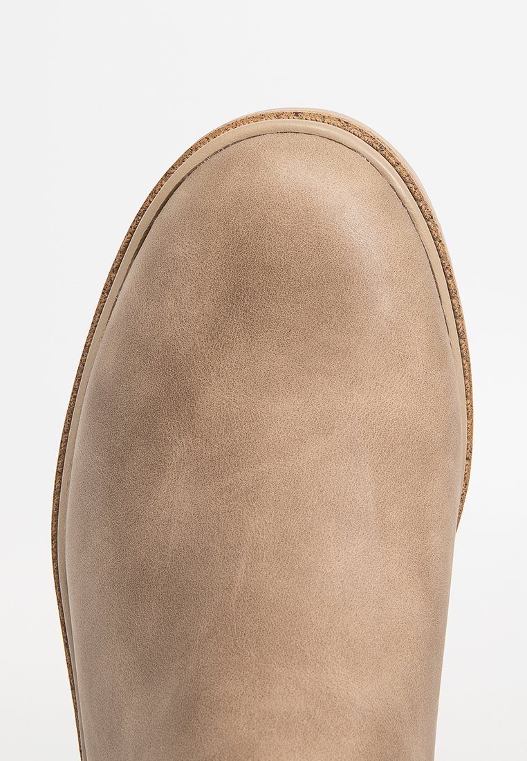 Nahaufnahme der abgerundeten Spitze eines beigen Wildlederschuhs mit sichtbaren Nähten und einem Kork-Geschmackrand.