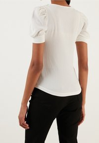 Biała bluzka o teksturze z krótkimi bufkami i dopasowanym krojem, noszona z czarnymi spodniami. Widok z tyłu ukazuje długość i fason ubrania.