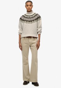 Maglione grigio chiaro a maglia con motivo a pois e collo alto, abbinato a pantaloni beige e scarpe marroni, in piedi su uno sfondo bianco.