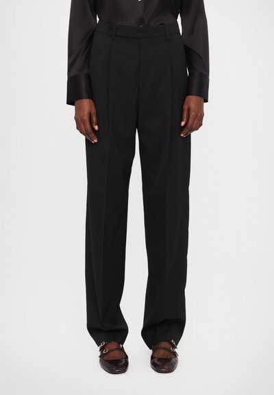 Schwarze, taillierte Hose mit hoher Taille, die zwei vordere Falten aufweist und aus einem glatten Stoff gefertigt ist. Getragen mit dunklen, strukturierten Schuhen.