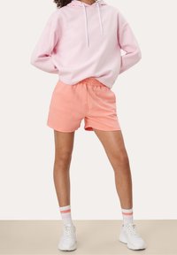Felpa rosa chiaro con cappuccio e cordini, abbinata a pantaloni corti corallo con vita elastica. Sneakers bianche con dettagli rosa.
