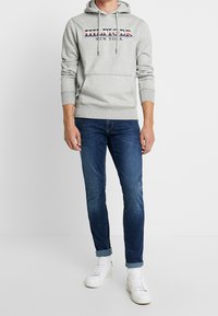 Sudadera con capucha gris con el logo "HILFIGER NEW YORK" en rojo y azul, combinada con jeans azul oscuro y zapatillas blancas. Corte casual.