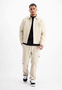 Beige canvas jas met drukknoopsluiting en borstzak, gecombineerd met bijpassende beige cargobroek met zijzakken en trekkoord, gedragen met zwarte sneakers.