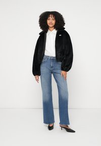 Γυναίκα με σγουρά μαλλιά που φορά μαύρο ζεστό μπουφάν, λευκό πουκάμισο με κουμπιά, μπλε τζιν με διαβάθμιση χρώματος και μαύρα mule με τακούνι, στέκεται μπροστά σε λευκό φόντο.