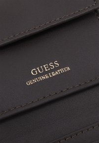 Superficie in pelle nera con cuciture fini e testo in rilievo oro con la scritta "GUESS GENUINE LEATHER" su una patta.