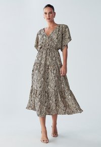 Rakstaina maksi kleita bēšā krāsā ar melnām viļņotām svītrām, ar V-veida izgriezumu, īsām platiem piedurknēm un saplacinātu vidukli, izgatavota no viegla auduma.