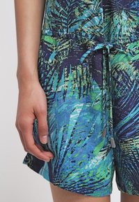 Main reposant à côté d’un short à motif feuilles tropicales bleu, vert et noir avec un cordon noué à la taille.
