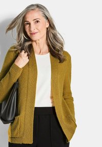Lächelnde Frau mittleren Alters mit grauem Haar, die eine senfgelbe Strickjacke, ein weißes Oberteil, schwarze Hosen trägt und eine schwarze Schultertasche vor einem weißen Hintergrund hält.