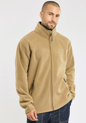 Homme portant une veste polaire beige zippée avec un col montant et des poches zippées, debout devant un fond clair uni.