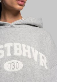 Felpa grigia con un grande logo bianco in peluche "STBHVR" e "03D" sul davanti. Presenta un cappuccio con cordino e un tessuto morbido e testurizzato.