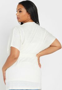 Vit stickad kappa med korta ärmar, V-ringning och matchande bälte. Texturerat, något genomskinligt tyg med en slät yta.