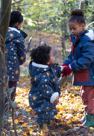 Drei Kinder in bunten Oberbekleidungen erkunden einen Wald. Eines trägt einen navyblauen Mantel mit himmlischen Mustern; die anderen haben unterschiedliche Farben und Texturen.