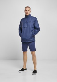 Mørkeblå letvægts halv-zip jakke med frontlomme, lange ærmer og elastiske manchetter, parret med mørkeblå shorts og sorte slip-on sko.