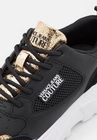 Svarta sneakers med en texturerad läderovansida med guldaccenter, nätpaneler, perforerade detaljer och en vit gummisula.