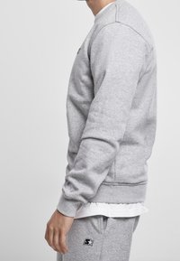 Sudadera gris con puños y dobladillo acanalados, que presenta un pequeño logo negro en el pecho. La tela parece suave y ligeramente texturizada.