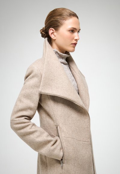 Beige uldblandingsfrakke med stor krave, lynlåslommer på siden, tætsiddende silhuet og blød tekstur. Båret over en lys grå sweater.