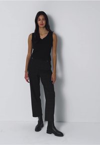 Musta ilmanhiha toppi V-kauluksella, yhdistettynä tummiin ruudullisiin korkeavyötäröisiin housuihin ja mustiin nilkkureihin, seisoo valkoista taustaa vasten.