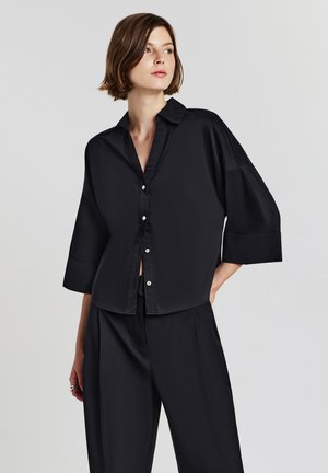 Woman wearing a loose black satin button-up shirt with three-quarter sleeves and matching wide-leg pants, standing against a plain background.