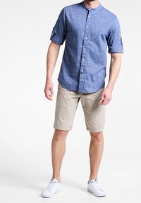 Mand iført en blå skjorte med opdragne ærmer, beige shorts og hvide sneakers, der står foran en hvid baggrund.