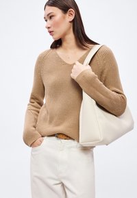 Beige strikkegenser med V-hals og ribbestruktur. Kombinert med kremfargede bukser og en hvit skinnveske på skulderen.