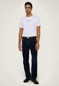 Bílé tričko s krátkým rukávem s logem "TRUSSARDI", v kombinaci s tmavě modrými džíny a černými botami. Jednoduchý design, pohodlný střih.