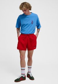 Blå t-shirt med kort ärm och grafiskt tryck, tillsammans med röda shorts med sidofickor och snörmidja. Gröna och röda sneakers.