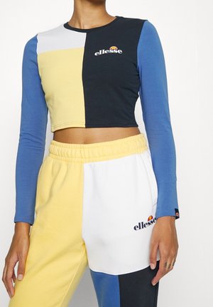 Modèle portant un ensemble crop top et pantalon de survêtement color-block en bleu, jaune, blanc et noir avec des logos "ellesse" sur le haut et le pantalon.