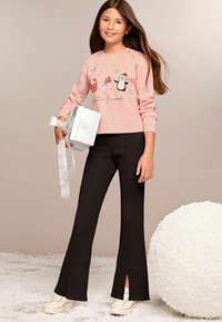 Sudadera rosa claro con gráfico de pingüino y texto, combinada con pantalones acampanados negros con canalé y aberturas, y zapatillas blancas. Sosteniendo un regalo.