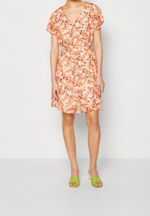 Vrouw draagt een korte mouw, V-hals bloemenjurk in oranje en roze tinten, gecombineerd met limegroene open teen sandalen met hak.