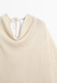 Pull beige en maille avec un col châle et de fines cordelettes à nouer à l'arrière, sur fond blanc.