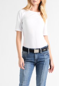 White short-sleeve top paired with blue denim jeans, featuring a black belt with a silver buckle and Lacoste logo accents.
