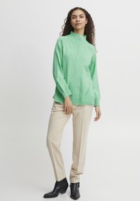 Pulover mint, cu mânecă lungă, guler înalt și fermoar pe jumătate. Textură cordoată la guler și la tiv. Asortat cu pantaloni bej, croiți.