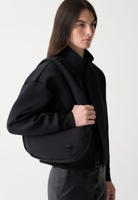 Fekete válltáska sima, párnázott textúrával, kerek logóval díszítve. Egy hozzá illő fekete cipzáros kabát és elegáns nadrág mellett viselve.