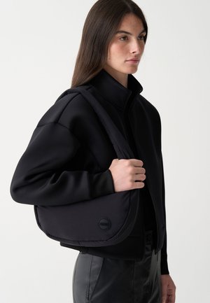 Fekete válltáska sima, párnázott textúrával, kerek logóval díszítve. Egy hozzá illő fekete cipzáros kabát és elegáns nadrág mellett viselve.
