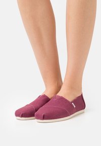 Burgundy canvas slip-on shoes with a rounded toe, contrasting light sole, and V-shaped cutouts on the sides for flexible fit.