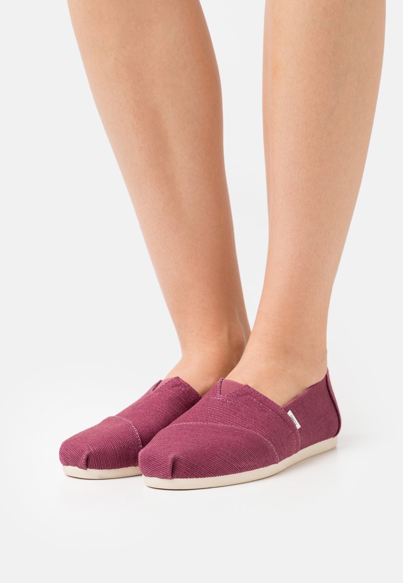 Burgundy canvas slip-on shoes with a rounded toe, contrasting light sole, and V-shaped cutouts on the sides for flexible fit.