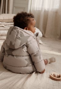 Veste matelassée grise avec de grands motifs d'étoiles, dotée d'une capuche et d'une doublure douce. Un enfant est assis sur une couverture beige texturée avec un jouet en bois.