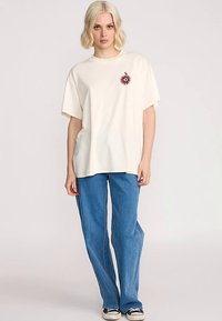 Cream cotton t-shirt with a floral embroidery design on the chest, paired with blue straight-leg jeans and black sneakers.