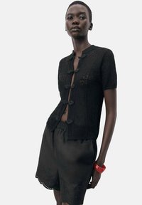 Schwarze gestrickte Strickjacke mit kurzen Ärmeln, versehen mit dekorativen Schlaufen als Verschlüsse. Kombiniert mit schwarzen Shorts und einem roten Armbandschmuck.