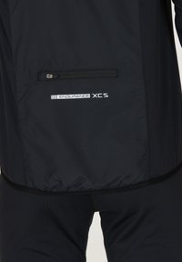 Sort atletisk jakke med en lynlåslomme på ryggen, struktureret stof, elastisk nederkant og hvid "ENDURANCE XCS" branding.