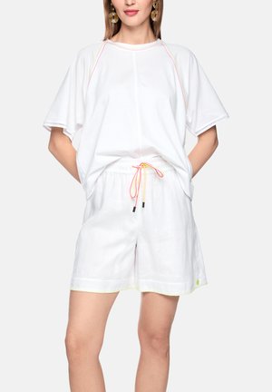 Frau trägt ein locker sitzendes weißes kurzärmeliges Oberteil und weiße Shorts mit gelbem und rosa Kordelzug, steht mit den Händen in den Taschen.